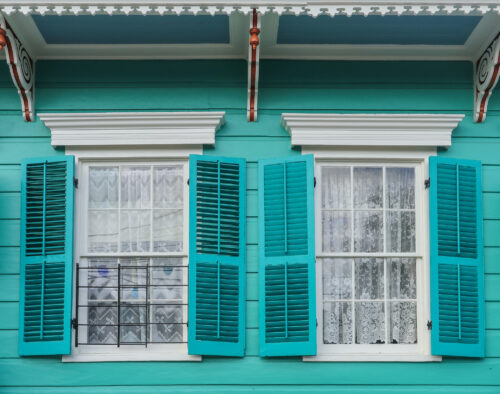 Close-up of a turquoise New Orleans house with two white-trimmed windows, open turquoise shutters, lace curtains, and decorative architectural details.