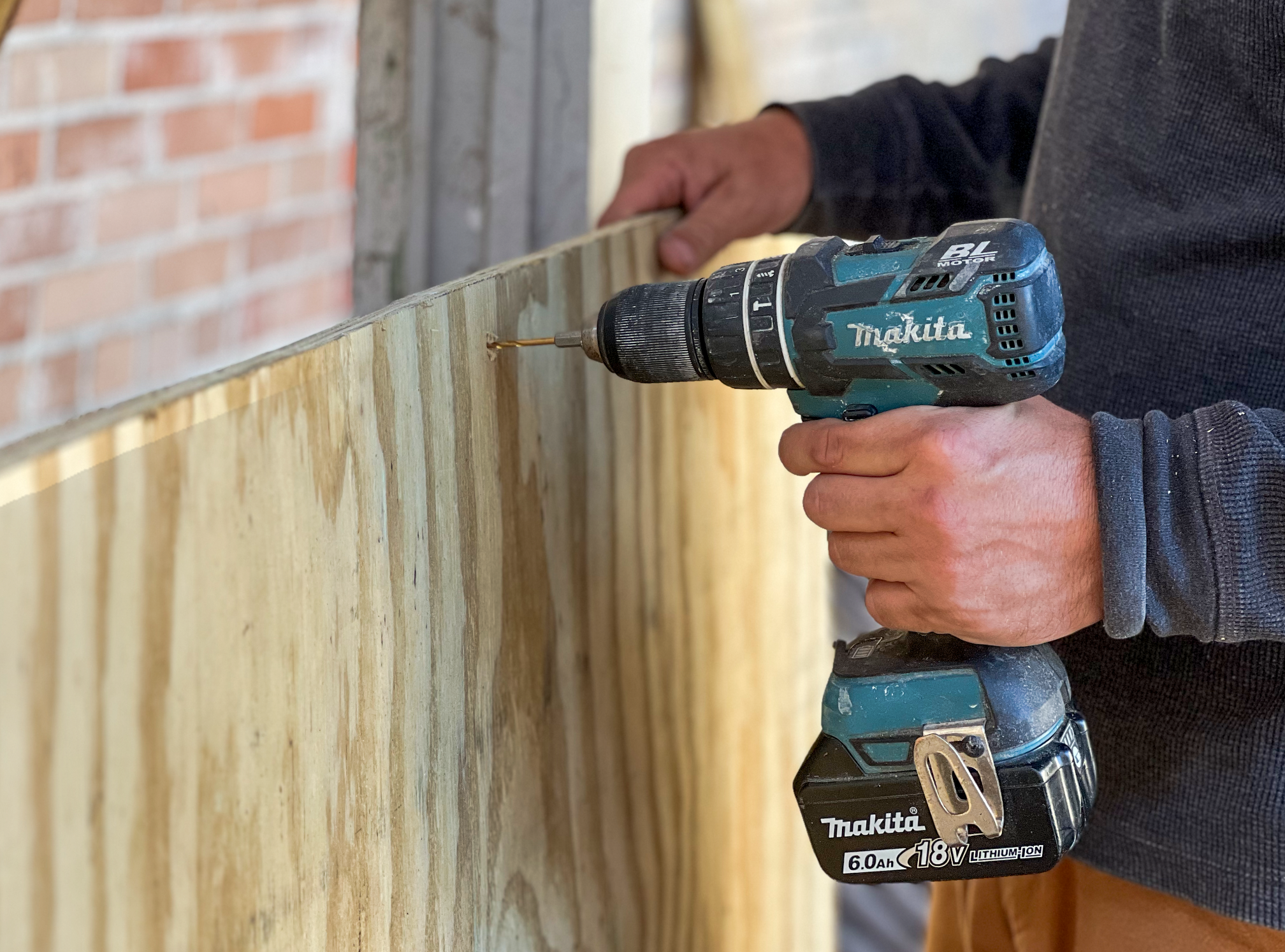 Close-up of a craftsperson using a cordless drill to fasten a screw into a sheet of plywood during a construction project. The image highlights hands-on preservation work, with the drill, wood panel, and partially exposed brick wall emphasizing skilled building and restoration techniques.
