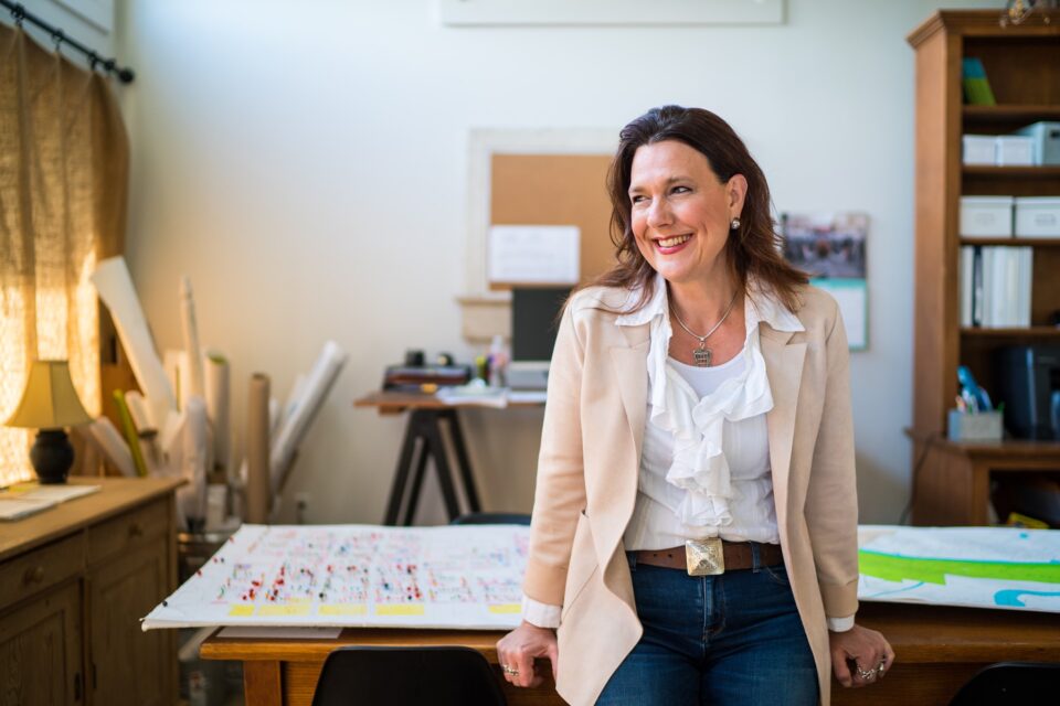 Kristin Gisleson Palmer smiles while standing in an office beside a table covered with architectural plans and drawings.