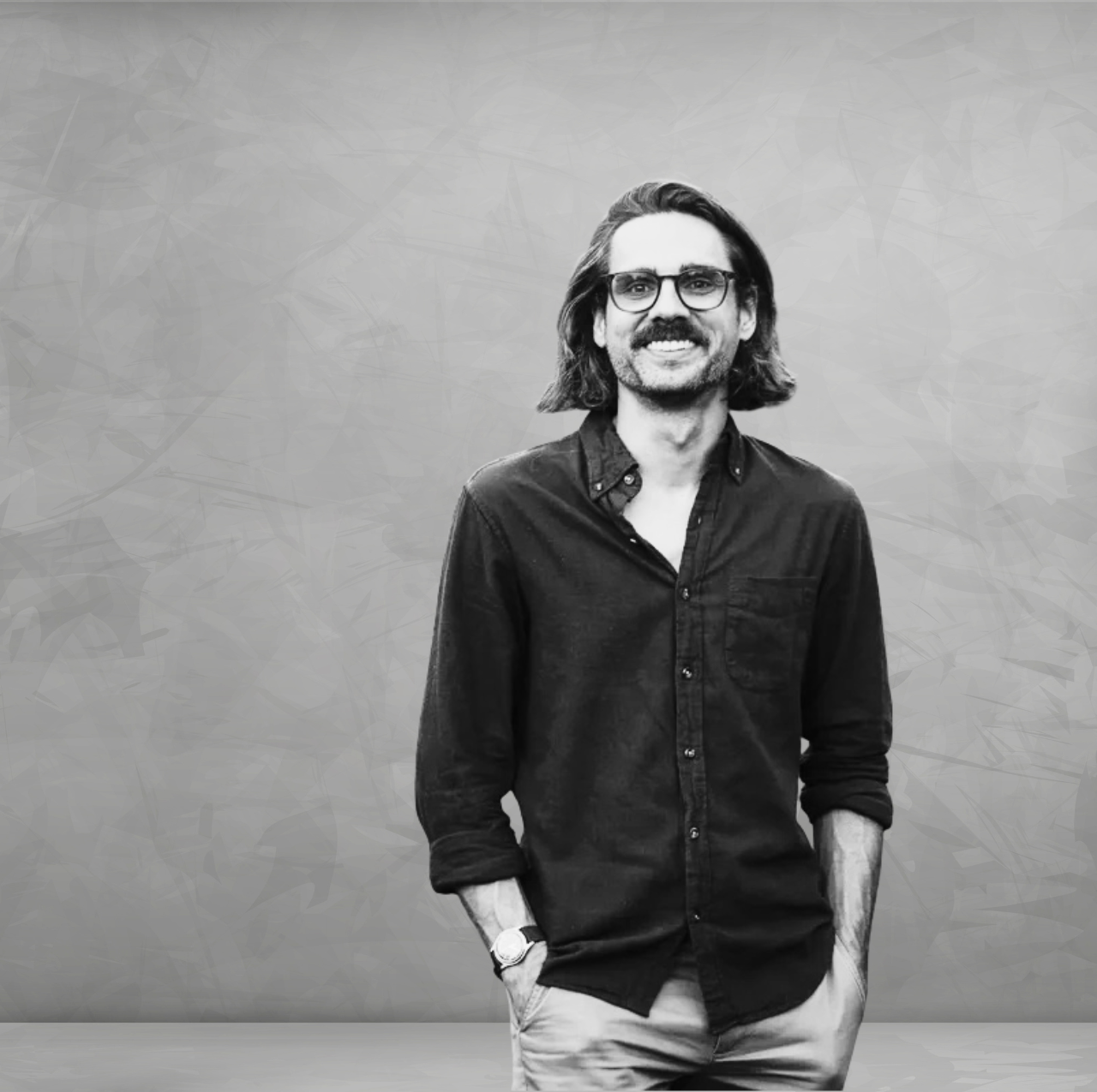 Black-and-white headshot of Alex Barthel, licensed architect, preservationist, and co-founder of Cultivo architecture firm. He is smiling and standing against a textured gray backdrop, wearing glasses, a dark button-down shirt with rolled sleeves, light-colored pants, and a wristwatch. His shoulder-length hair and relaxed stance with hands in his pockets create an approachable, professional portrait.