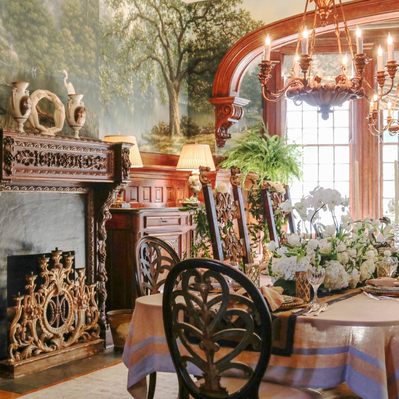 Ornate formal dining room with carved dark woodwork, a mural-covered wall, elaborate chandeliers, and a table set with white flowers, fine china, and crystal glassware beside a decorative fireplace.