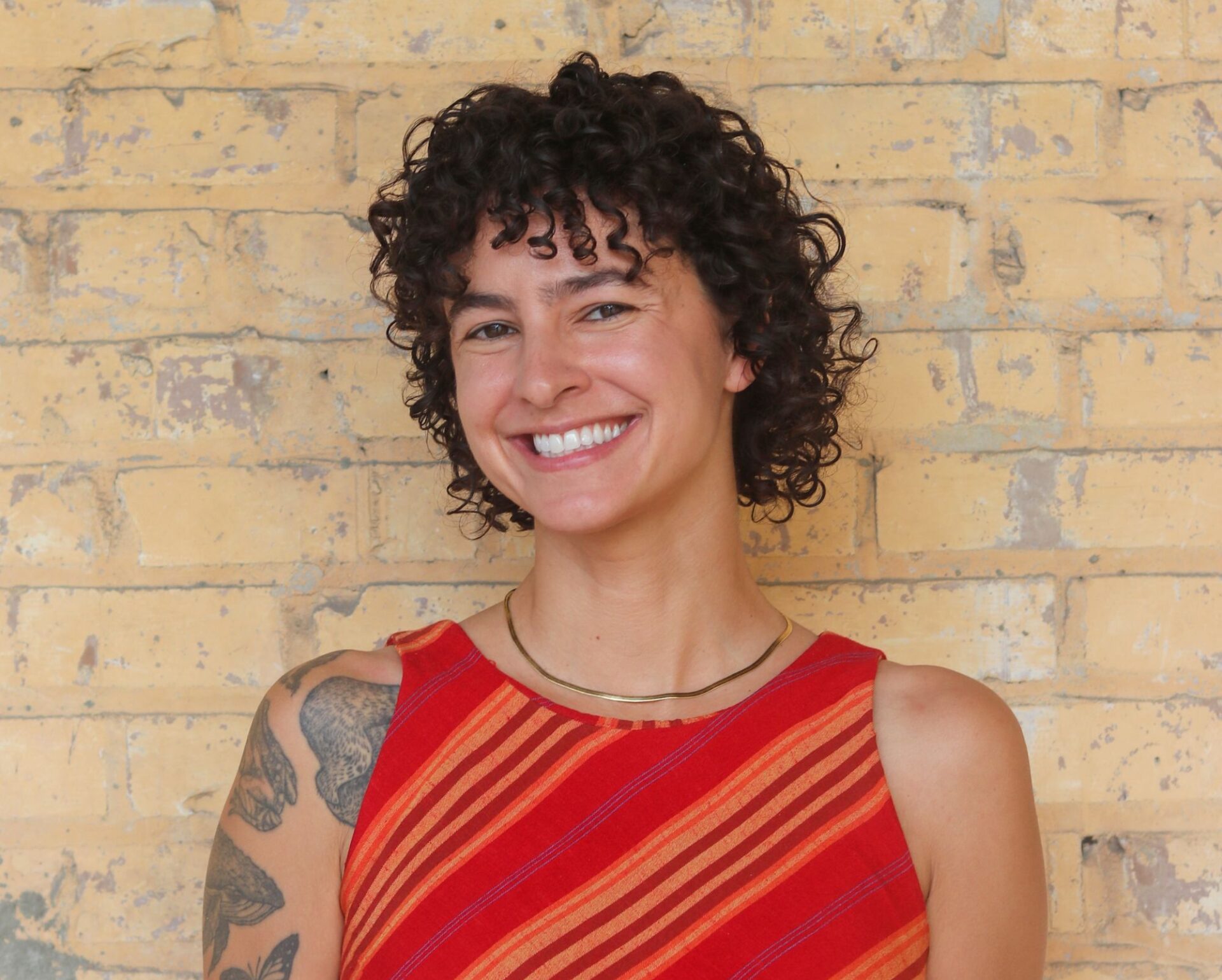 Portrait of Annie Agnone smiling in front of a light yellow brick wall. She has short dark curly hair and wears a sleeveless red-and-orange striped top, a gold necklace, and has a tattoo visible on her left shoulder.