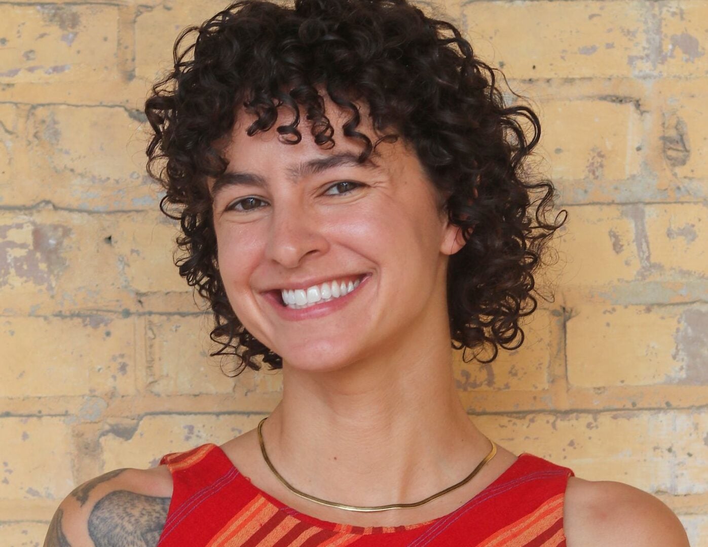 Portrait of Annie Agnone smiling in front of a light yellow brick wall. She has short dark curly hair and wears a sleeveless red-and-orange striped top, a gold necklace, and has a tattoo visible on her left shoulder.