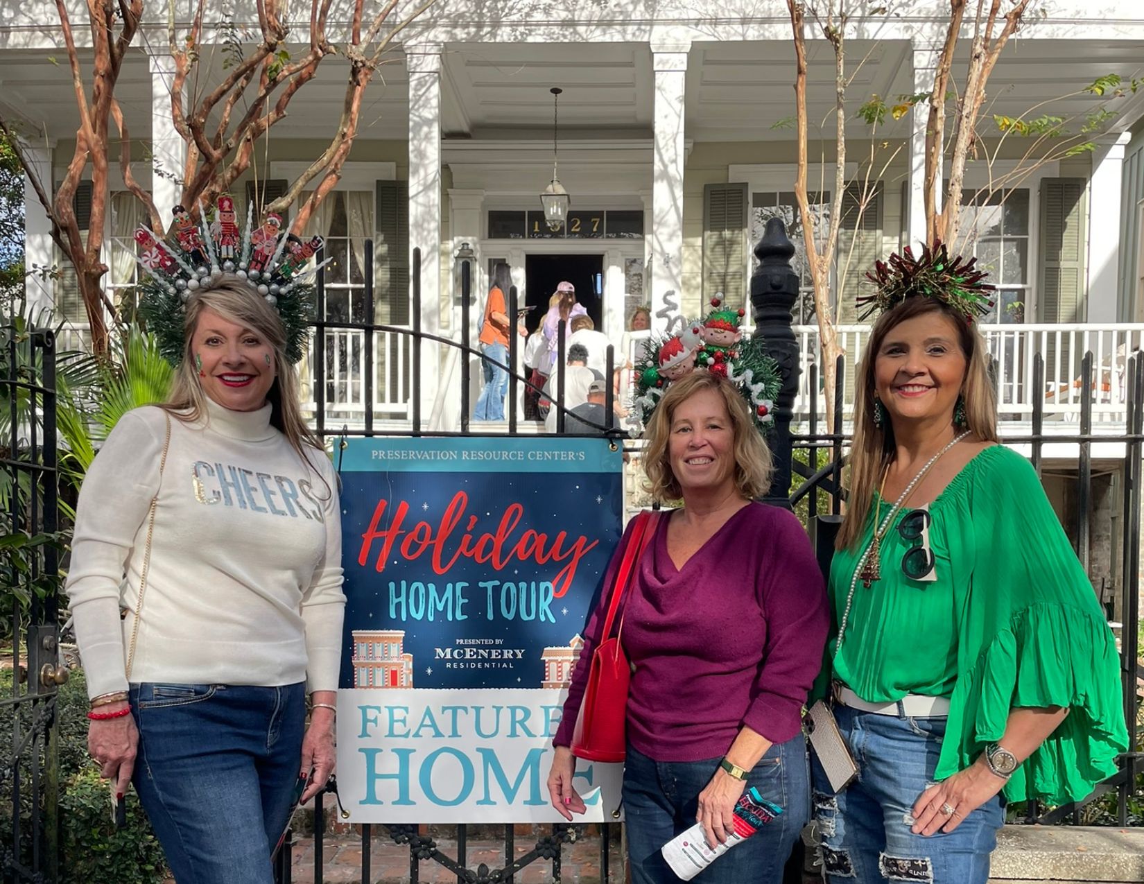 Three Holiday Home Tour attendees wearing festive holiday headbands pose beside a Holiday Home Tour sign in front of a featured historic New Orleans home. The decorated entrance and welcoming front porch provide a cheerful backdrop for the seasonal event.