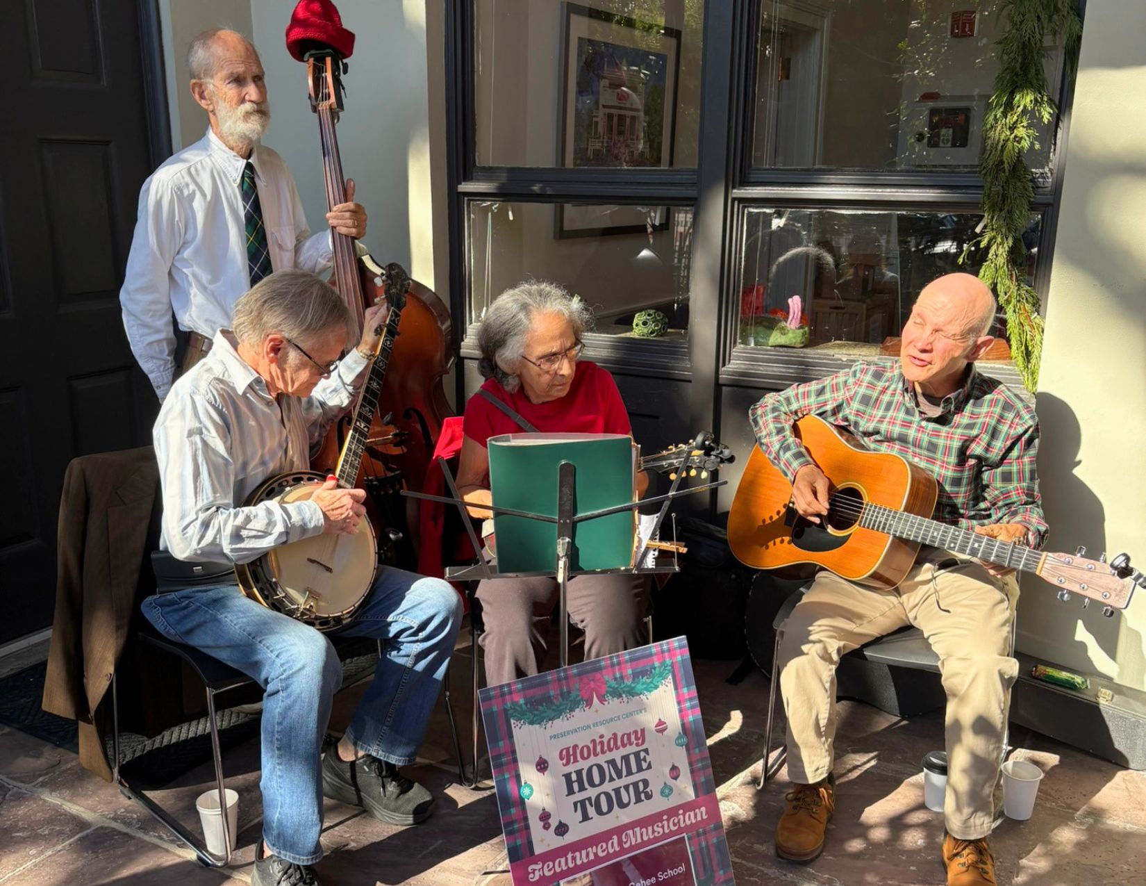 A four-piece acoustic ensemble performs on the porch of a Holiday Home Tour residence. The musicians play banjo, upright bass, mandolin, and acoustic guitar beside a sign reading "Holiday Home Tour Featured Musician," creating a festive atmosphere for tour guests.