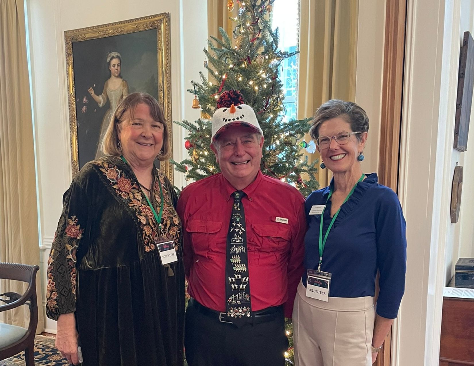 Three Holiday Home Tour volunteers smile in front of a decorated Christmas tree inside a historic home. The volunteer in the center wears a festive snowman hat, red shirt, and holiday tie, while the two volunteers beside him wear name badges and stand beneath a framed portrait in the elegant, holiday-decorated interior.