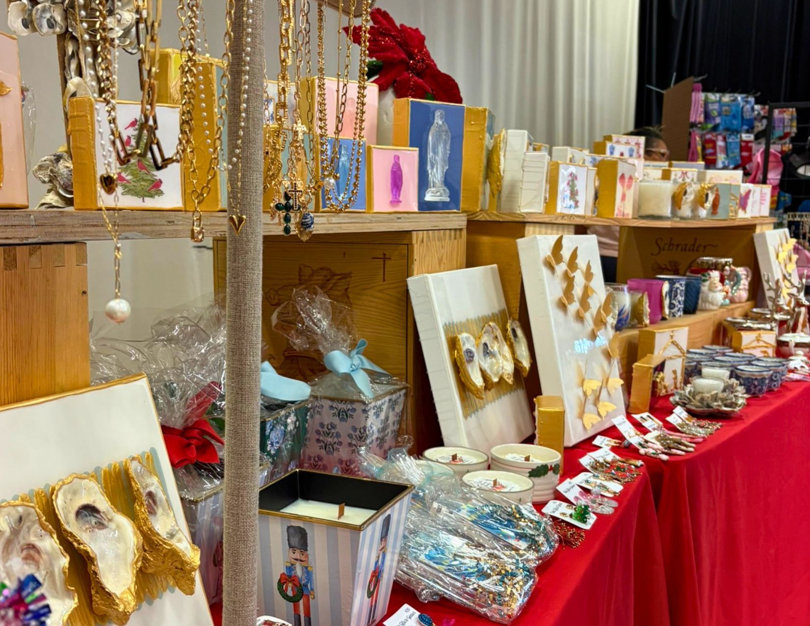 A holiday market vendor booth displays handmade jewelry, candles, artwork, ornaments, and gift items arranged across red tablecloth-covered tables. Gold-accented oyster shell art, necklaces, earrings, and festive décor create a colorful holiday shopping display.