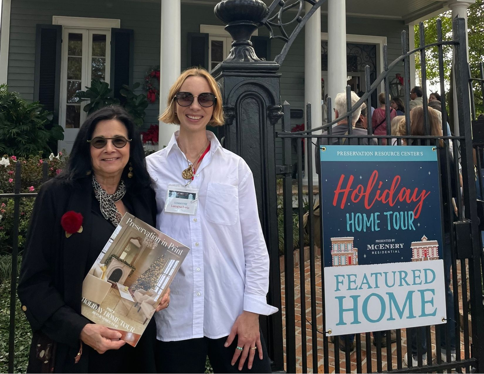 Two Holiday Home Tour attendees pose beside a Holiday Home Tour Featured Home sign outside a historic New Orleans residence. One attendee holds a copy of Preservation in Print featuring the Holiday Home Tour cover, while guests enter the decorated home in the background.