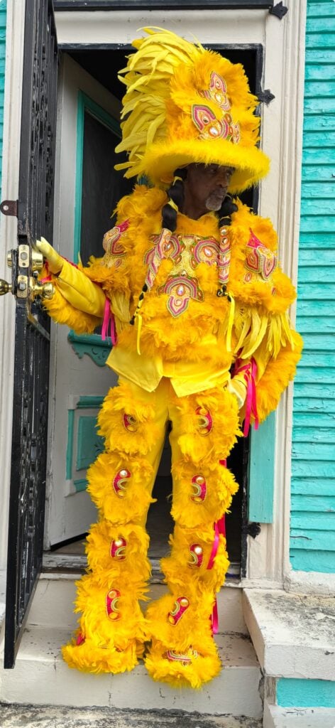Mardi Gras Indian in an elaborate yellow feathered suit and headdress standing in the doorway of a turquoise New Orleans house.