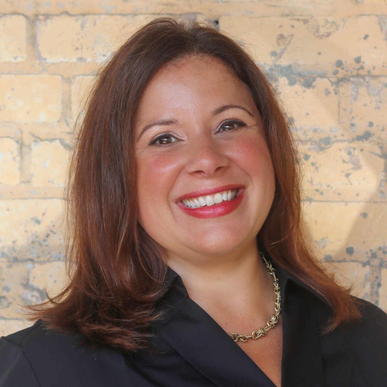 Headshot of Kristen Brooks, Director of Advancement for the Preservation Resource Center of New Orleans. She is smiling and wearing a black blouse and gold necklace, standing in front of a light tan brick wall.