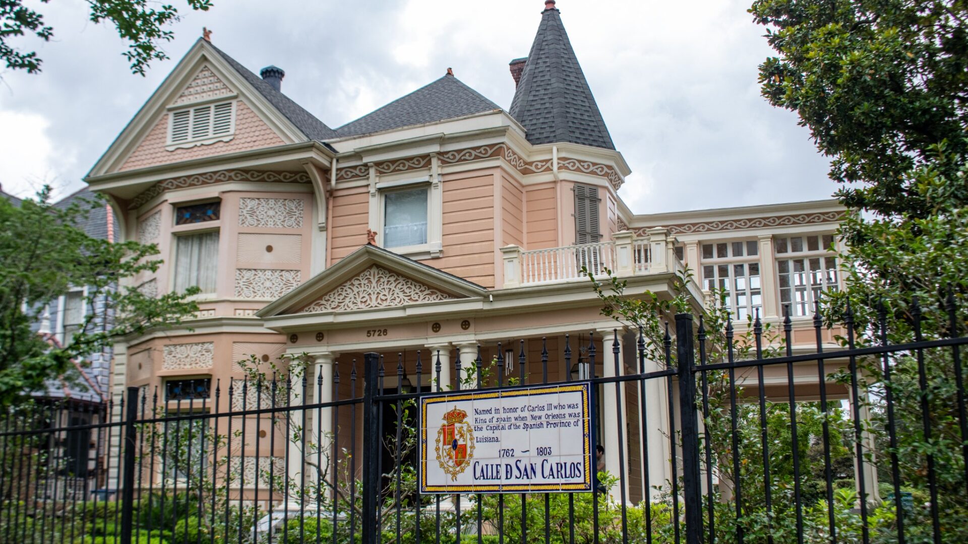 Historic Victorian home with intricate gingerbread trim, a corner turret, and wraparound porch behind an iron fence in New Orleans, with a "Calle de San Carlos" plaque in the foreground.