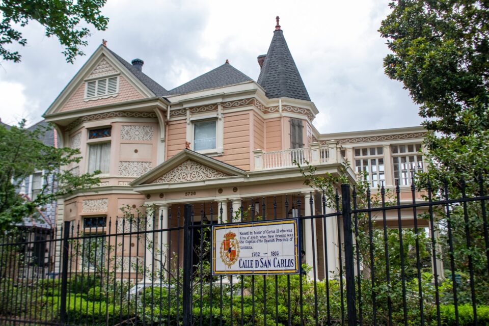 Historic Victorian home with intricate gingerbread trim, a corner turret, and wraparound porch behind an iron fence in New Orleans, with a "Calle de San Carlos" plaque in the foreground.