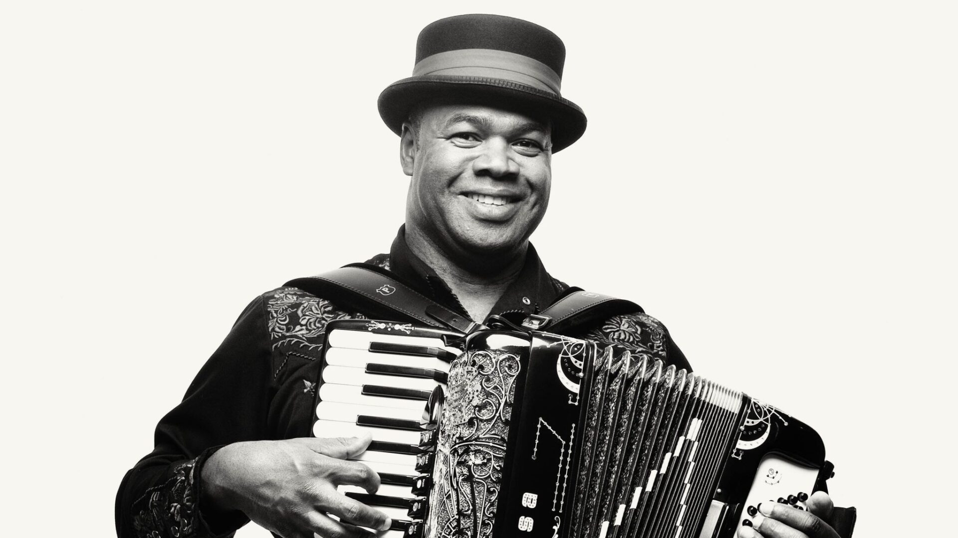 Black-and-white portrait of a smiling accordion player wearing a brimmed hat and embroidered shirt while holding and playing an accordion.