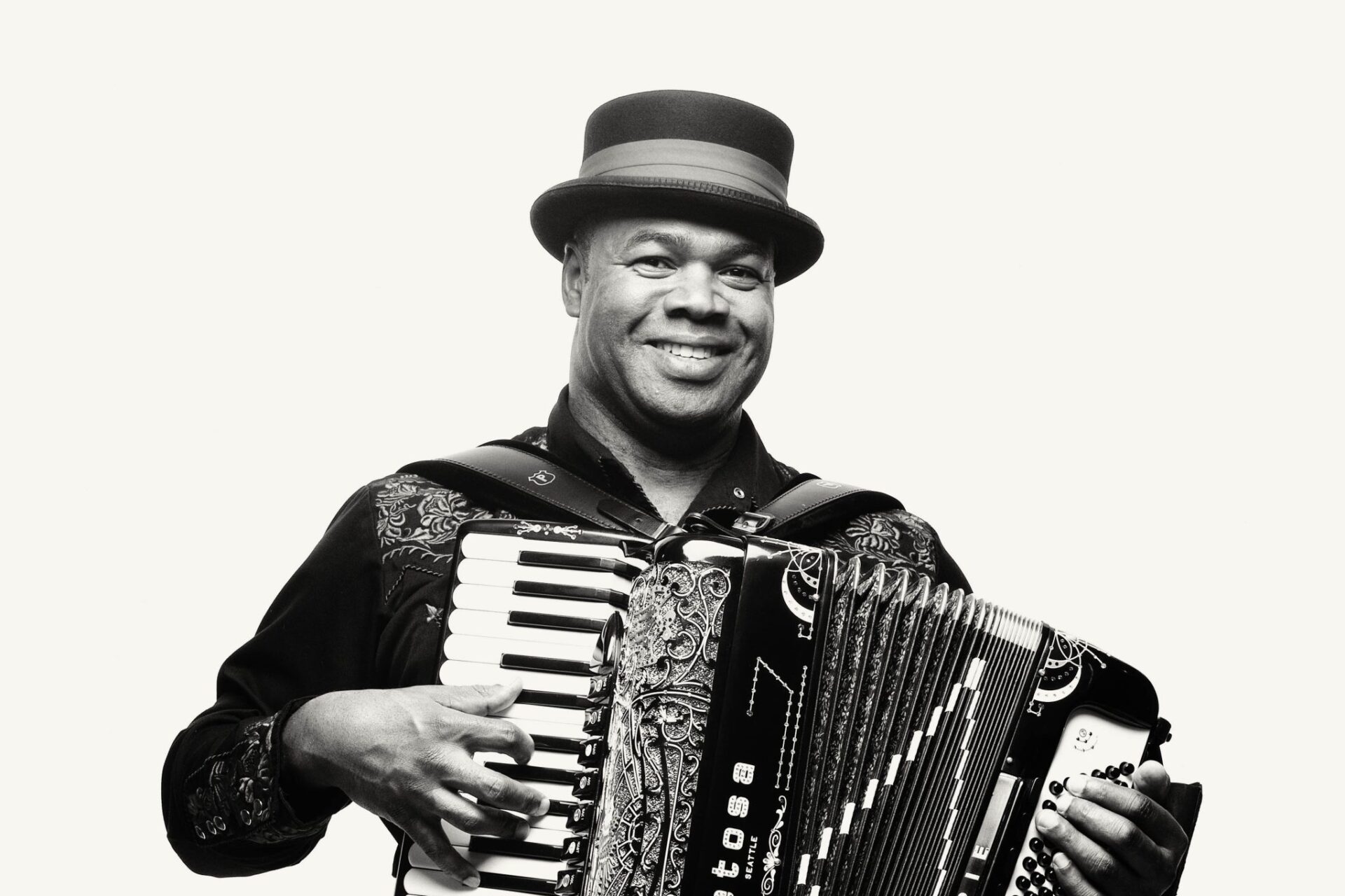Black-and-white portrait of a smiling accordion player wearing a brimmed hat and embroidered shirt while holding and playing an accordion.