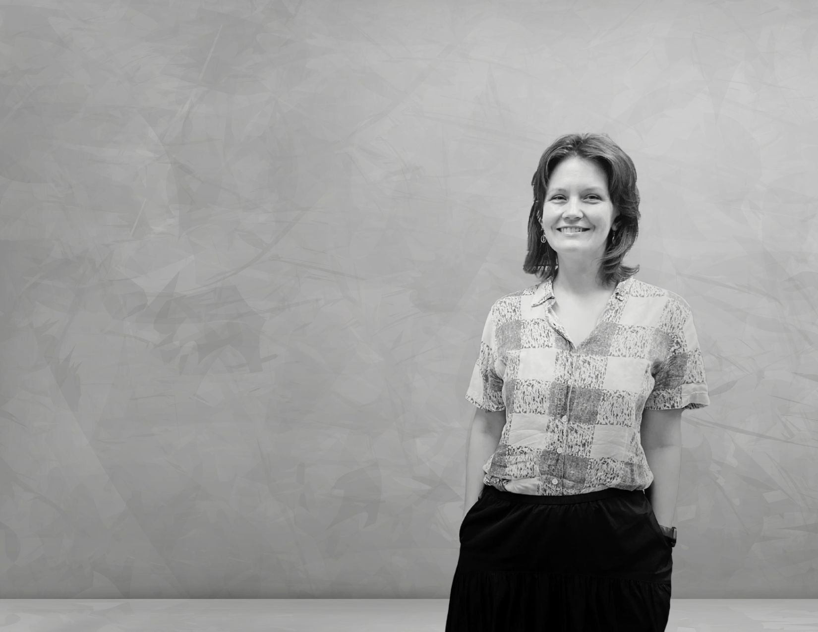 Black-and-white portrait of Amanda Fallis smiling and standing against a textured gray backdrop, wearing a patterned short-sleeve blouse and dark skirt.