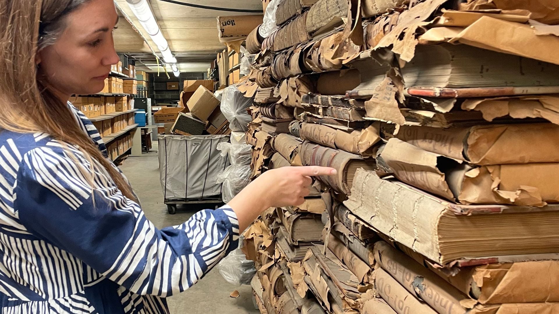 A woman examines and points to a stack of old, deteriorating bound documents and records in an archival storage area.