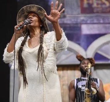 Singer Charmaine Neville performs onstage, singing into a microphone and raising one hand, with a musician playing saxophone behind her.