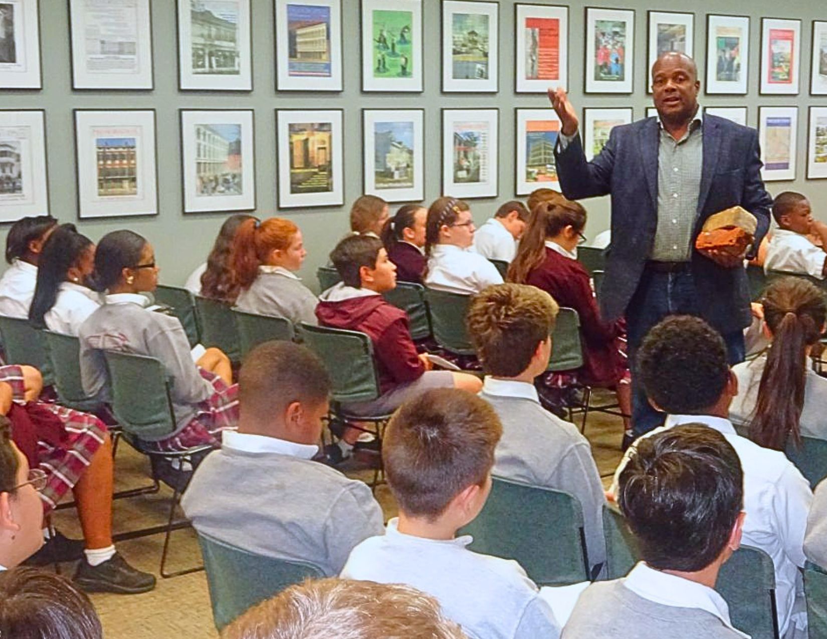 Students listen as a speaker addresses a seated group during an educational presentation, surrounded by framed artwork and photographs on the gallery walls.