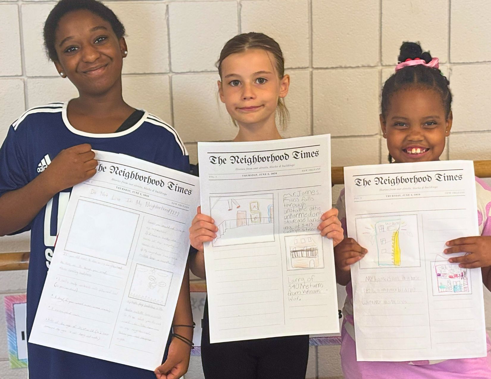 Three elementary school students proudly hold completed Neighborhood Times worksheets featuring drawings and written observations about their local community.
