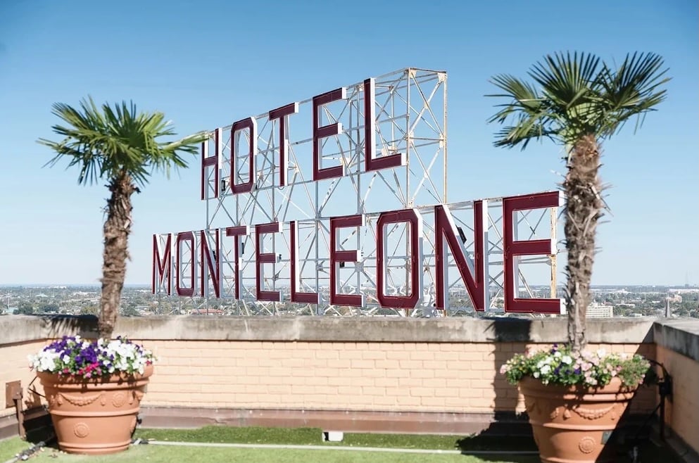 The rooftop sign of Hotel Monteleone in New Orleans, framed by palm trees and flower planters against a clear blue sky.