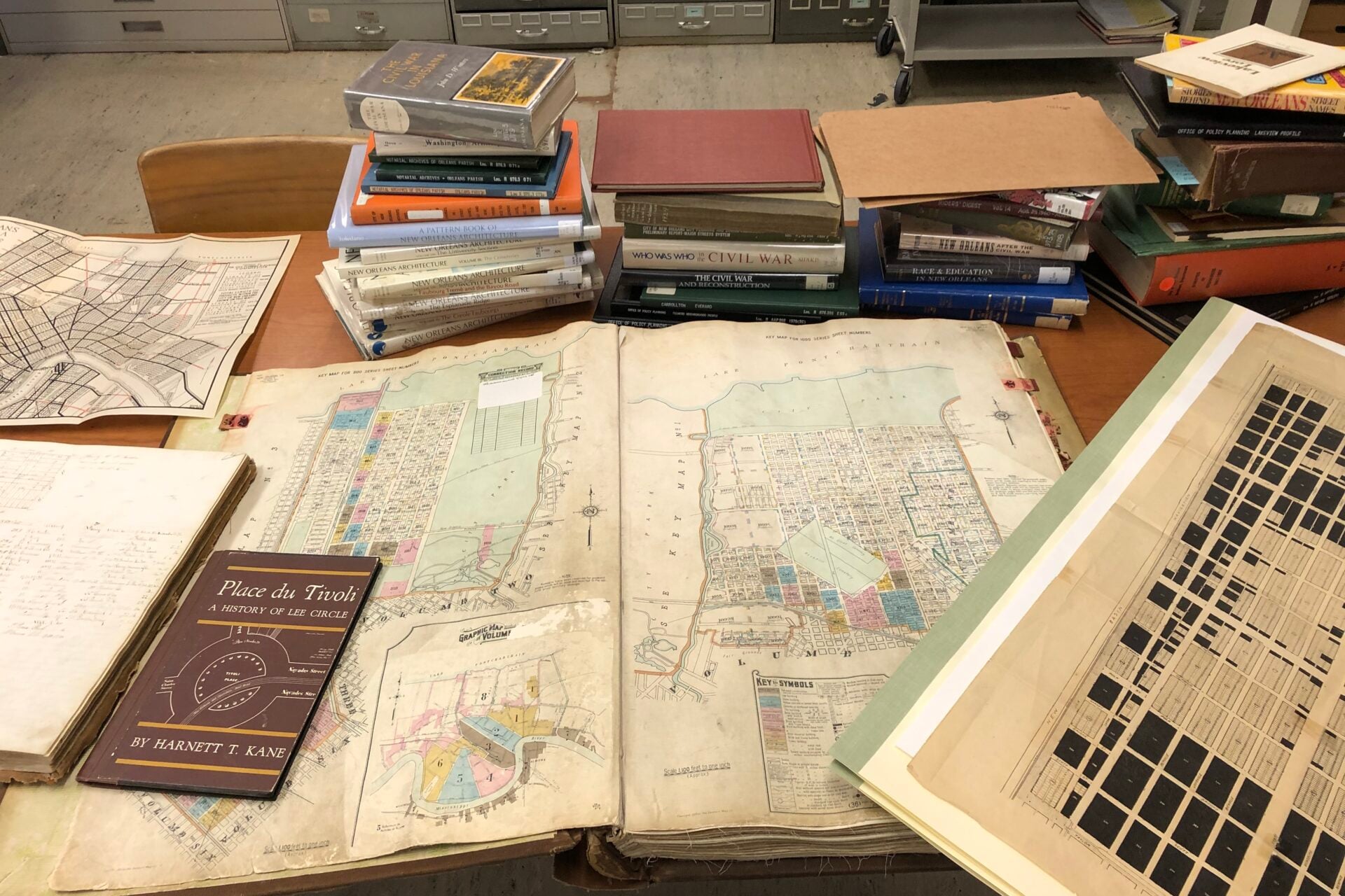 A research table covered with historic maps, books, architectural publications, and archival documents related to New Orleans. An open map dominates the center of the table, surrounded by stacks of books, city plans, and other historical materials.