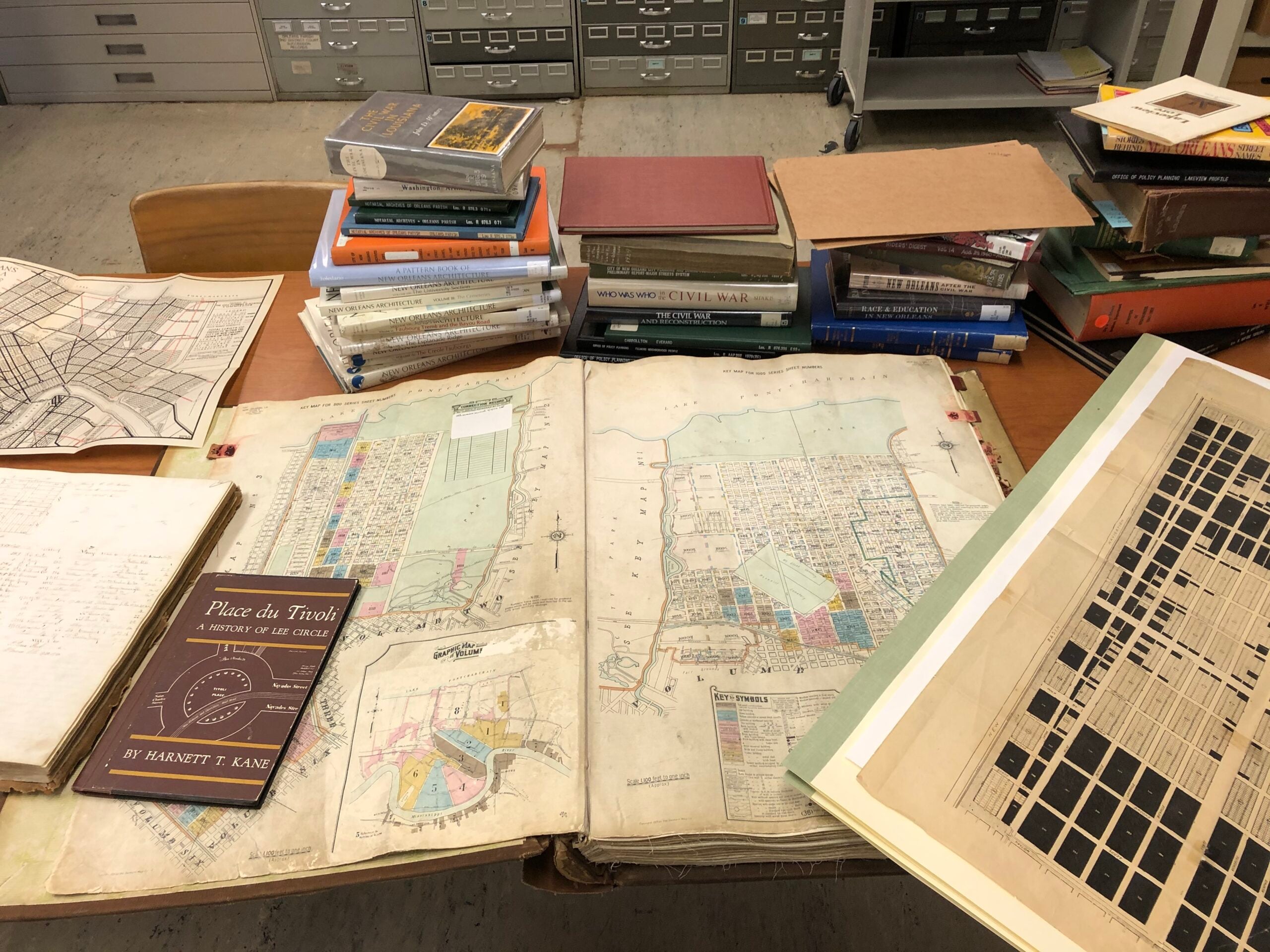 A research table covered with historic maps, books, architectural publications, and archival documents related to New Orleans. An open map dominates the center of the table, surrounded by stacks of books, city plans, and other historical materials.