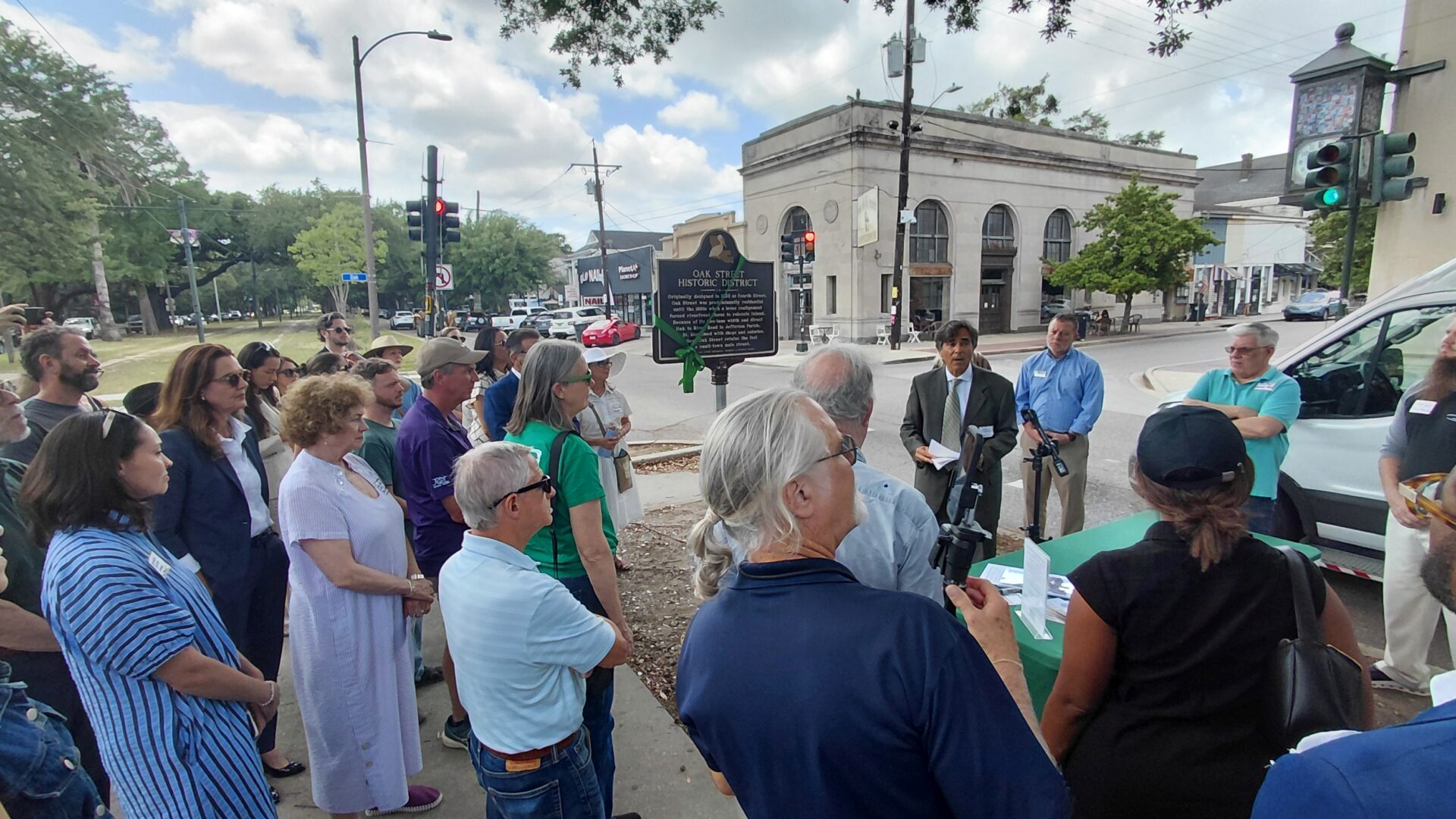 A crowd gathers at the corner of Oak Street and Carrollton Avenue for the dedication of the new Oak Street Historic District plaque. A speaker addresses attendees beside the marker while neighbors, preservation advocates, and community members look on. The historic commercial corridor and surrounding buildings frame the event, celebrating the neighborhood's architectural and cultural heritage. Photo by Richard Campanella.