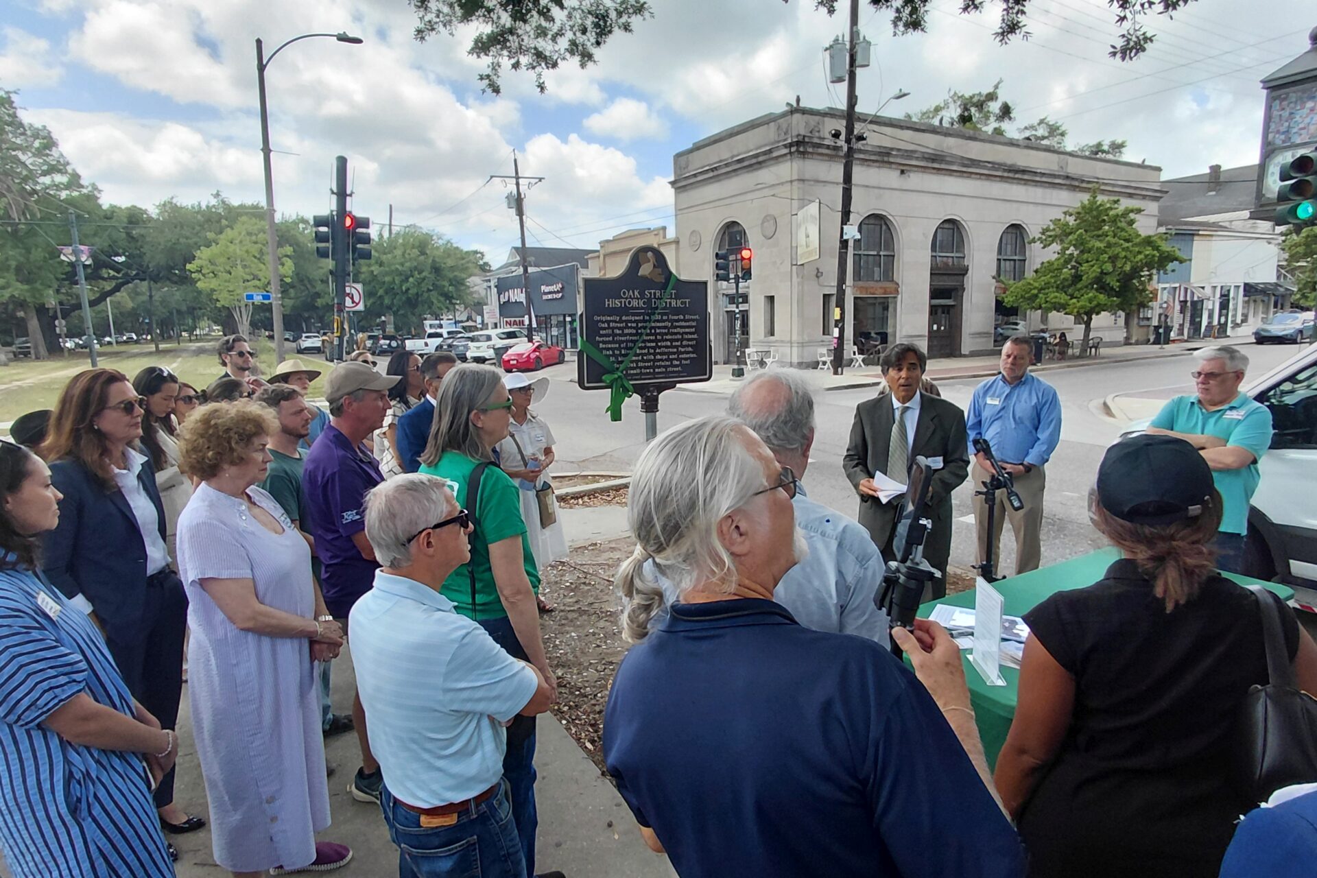 A crowd gathers at the corner of Oak Street and Carrollton Avenue for the dedication of the new Oak Street Historic District plaque. A speaker addresses attendees beside the marker while neighbors, preservation advocates, and community members look on. The historic commercial corridor and surrounding buildings frame the event, celebrating the neighborhood's architectural and cultural heritage. Photo by Richard Campanella.
