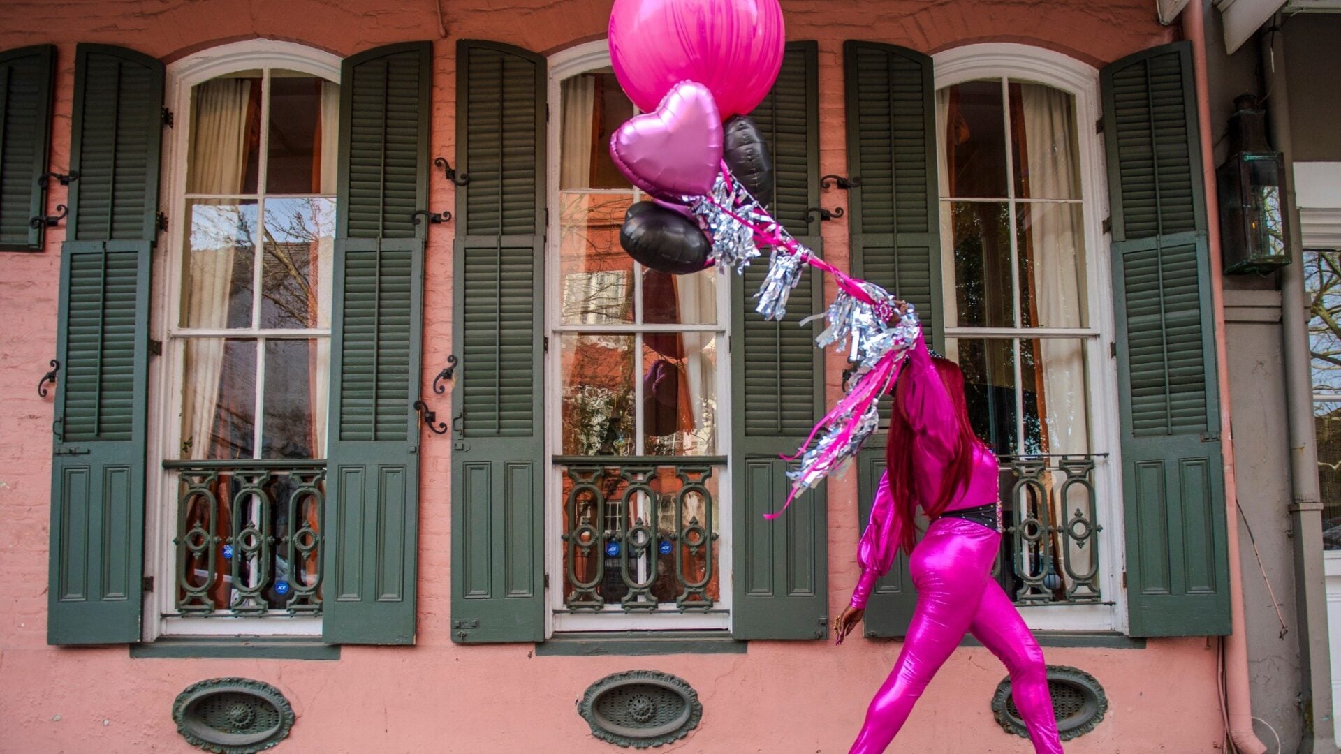 A performer in a bright pink outfit walks past a historic New Orleans home with pink and black balloons, framed by tall windows with dark green shutters.