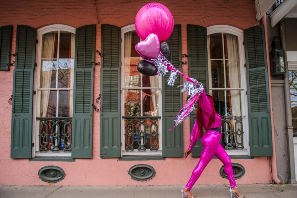 A performer in a bright pink outfit walks past a historic New Orleans home with pink and black balloons, framed by tall windows with dark green shutters.