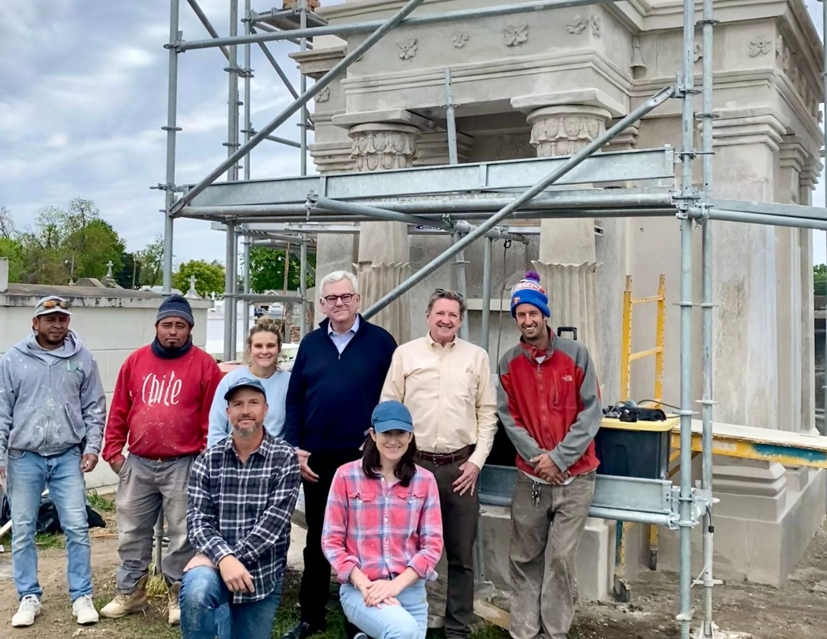 Conservation crew members pose with restorers Dennis Alonzo and Nathan Chapman in front of a historic cemetery tomb undergoing restoration, surrounded by scaffolding.