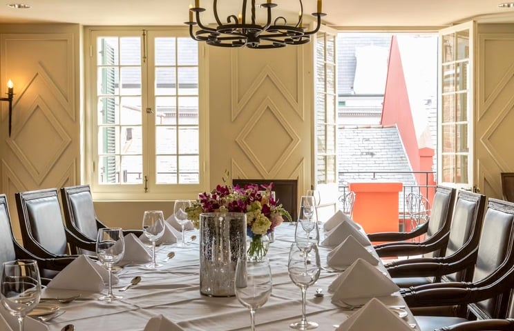Elegantly set dining table in the Tableau Parlor Room, with white linens, folded napkins, floral centerpiece, and views of New Orleans architecture through open windows.