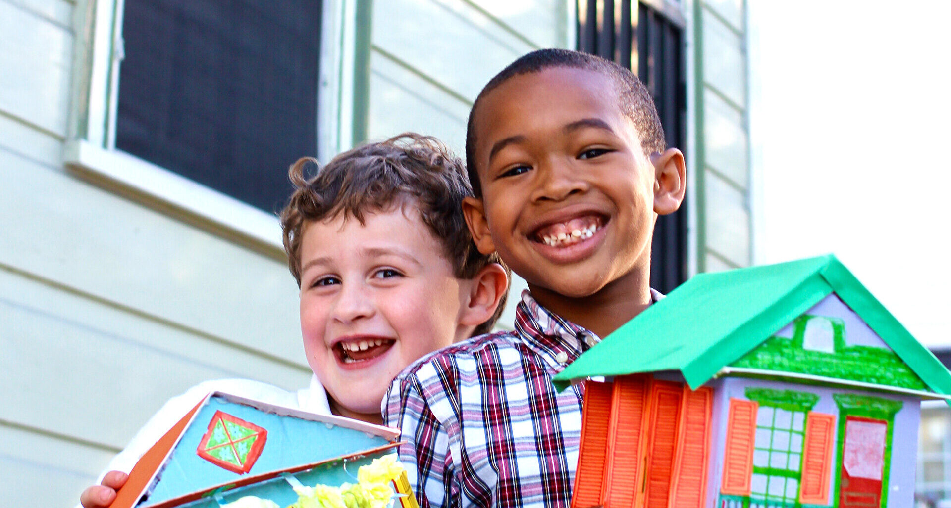 Two elementary school students smile while holding colorful handmade model houses during a neighborhood architecture learning activity.