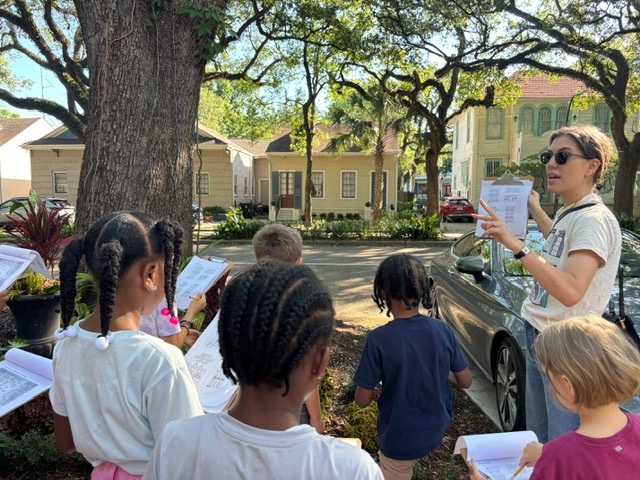 Students learn about historic New Orleans architecture during an outdoor neighborhood tour, listening to a teacher and taking notes while observing local homes.