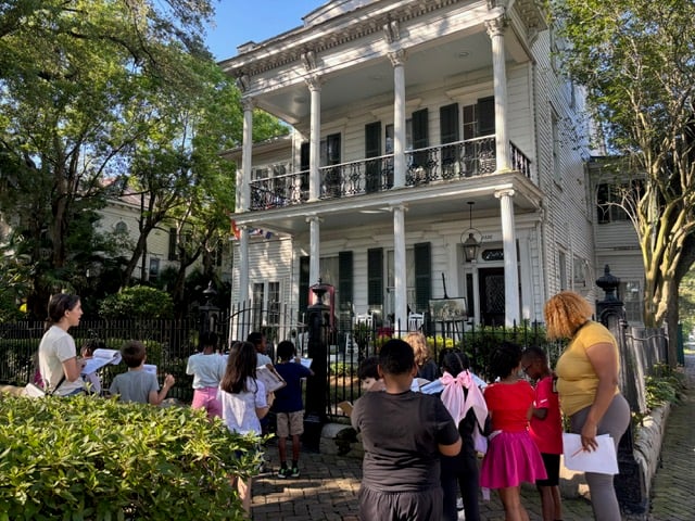 Elementary school students participate in a neighborhood learning activity outside a historic New Orleans home, observing the architecture and taking notes with their teachers.