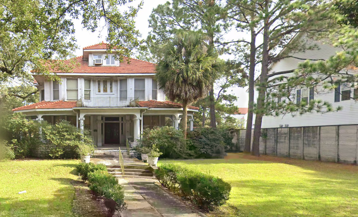 8133 Spruce Street, a historic two-story residence with a red tile roof, white siding, covered front porch, and second-story balcony, surrounded by mature trees and a landscaped lawn.