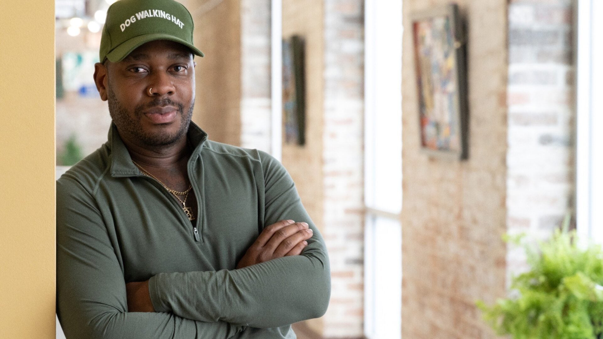 Dustin Burks stands with his arms crossed inside his home, wearing a green cap reading “Dog Walking Hat” and a matching green pullover. Exposed brick walls, large windows, artwork, and houseplants are visible in the background.