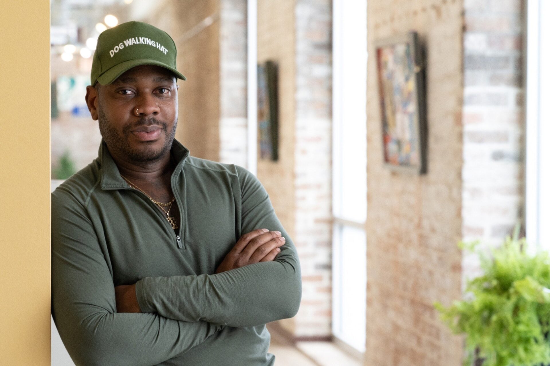 Dustin Burks stands with his arms crossed inside his home, wearing a green cap reading “Dog Walking Hat” and a matching green pullover. Exposed brick walls, large windows, artwork, and houseplants are visible in the background.
