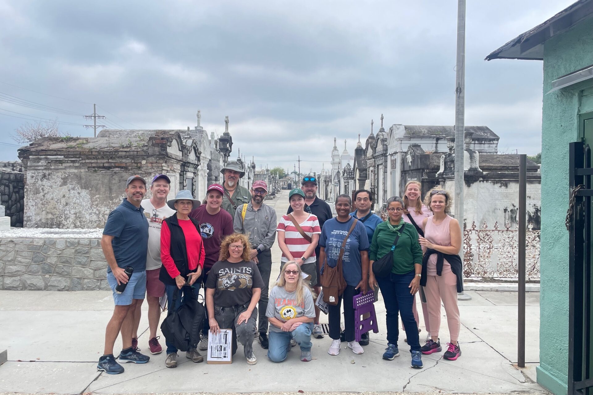 A group of people poses together during a cemetery tour, surrounded by rows of historic above-ground tombs and mausoleums under an overcast sky.