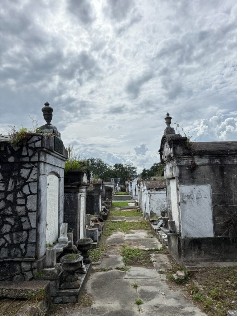 A narrow pathway through Lafayette Cemetery No. 1 lined with weathered above-ground tombs and mausoleums, some topped with decorative urns. Overgrown vegetation borders the cracked walkway beneath a dramatic cloudy sky.