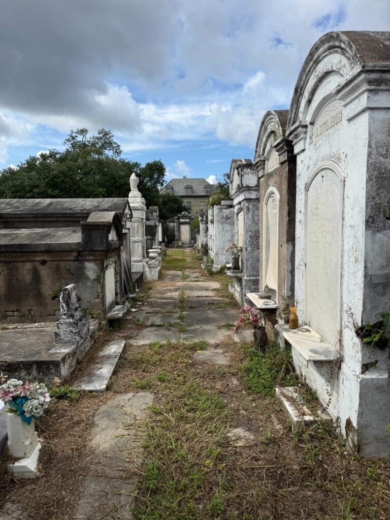 A narrow pathway through Lafayette Cemetery No. 1 lined with weathered above-ground tombs and mausoleums, including ornate arched memorials on the right. Cracked pavement and overgrown grass run between the tombs, with a historic house visible in the distance beneath a partly cloudy sky.