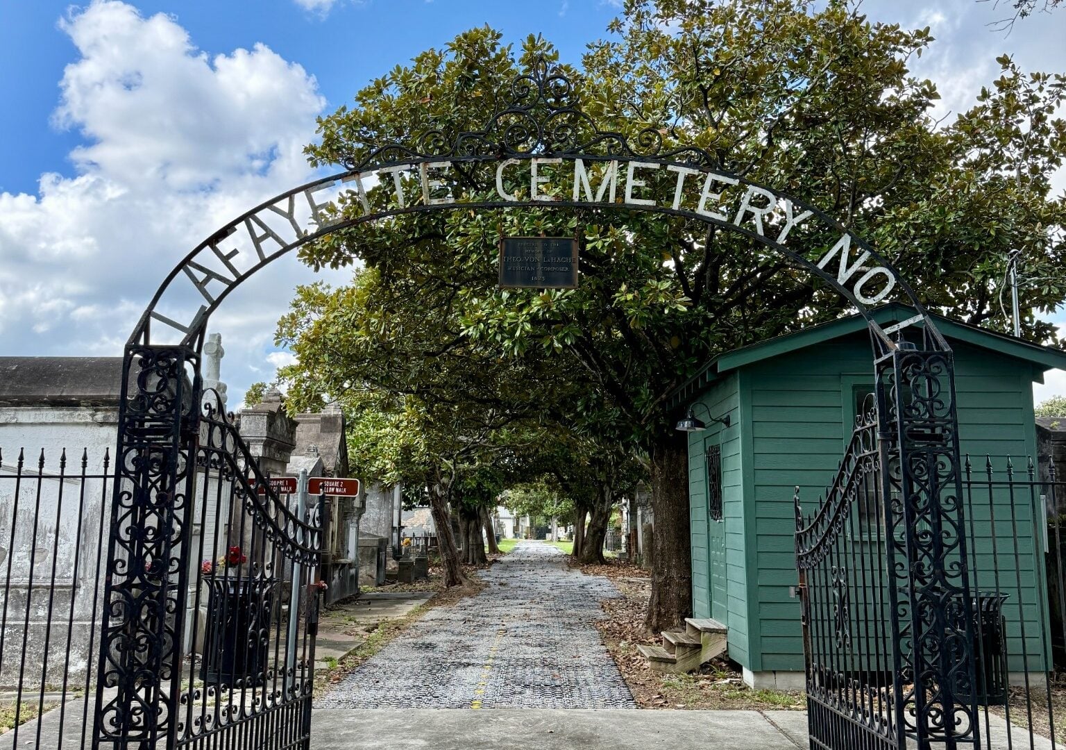 Ornate wrought-iron gates at the entrance to Lafayette Cemetery No. 1, with the cemetery name displayed across the arched gate. A tree-lined pathway and historic tombs extend into the cemetery, with a small green gatehouse on the right.