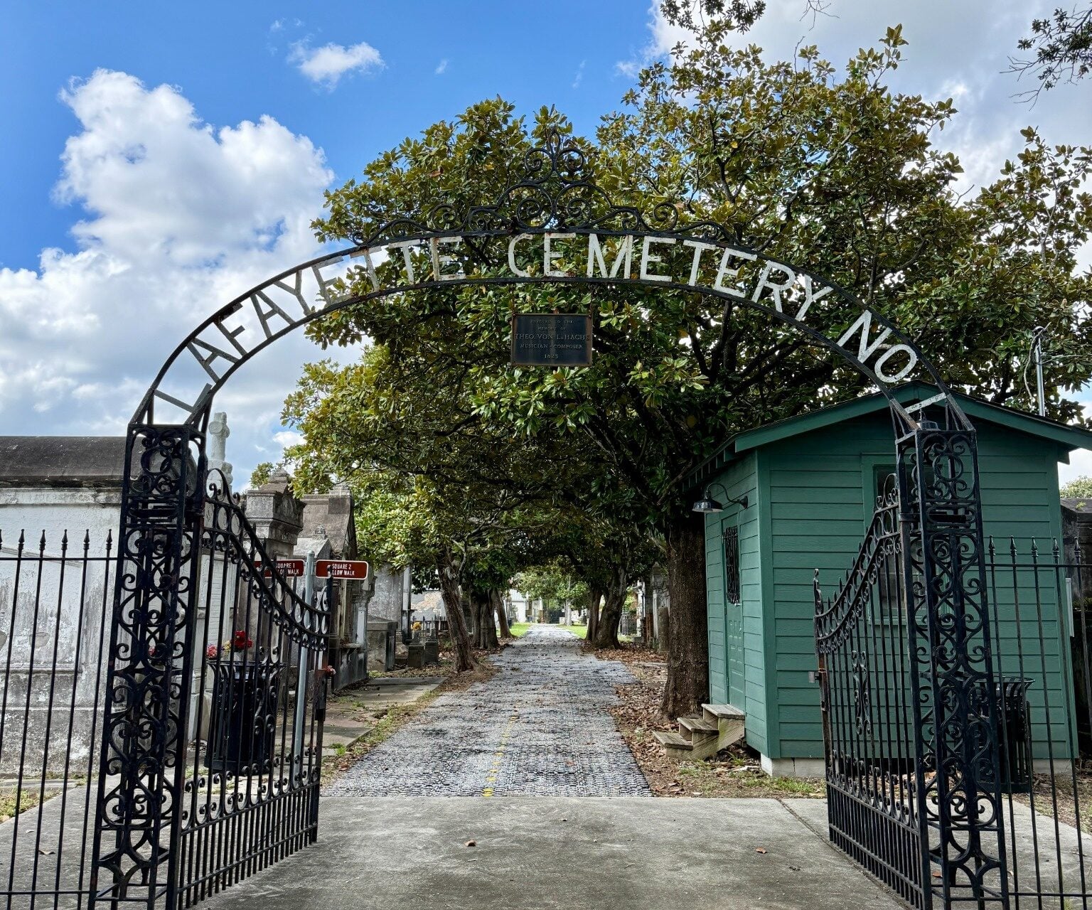 Ornate wrought-iron gates at the entrance to Lafayette Cemetery No. 1, with the cemetery name displayed across the arched gate. A tree-lined pathway and historic tombs extend into the cemetery, with a small green gatehouse on the right.