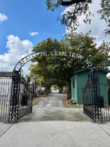 Ornate wrought-iron gates at the entrance to Lafayette Cemetery No. 1, with the cemetery name displayed across the arched gate. A tree-lined pathway and historic tombs extend into the cemetery, with a small green gatehouse on the right.