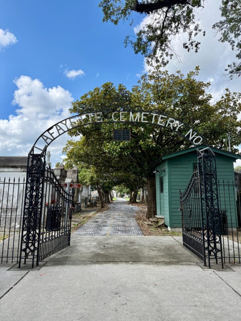Ornate wrought-iron gates at the entrance to Lafayette Cemetery No. 1, with the cemetery name displayed across the arched gate. A tree-lined pathway and historic tombs extend into the cemetery, with a small green gatehouse on the right.