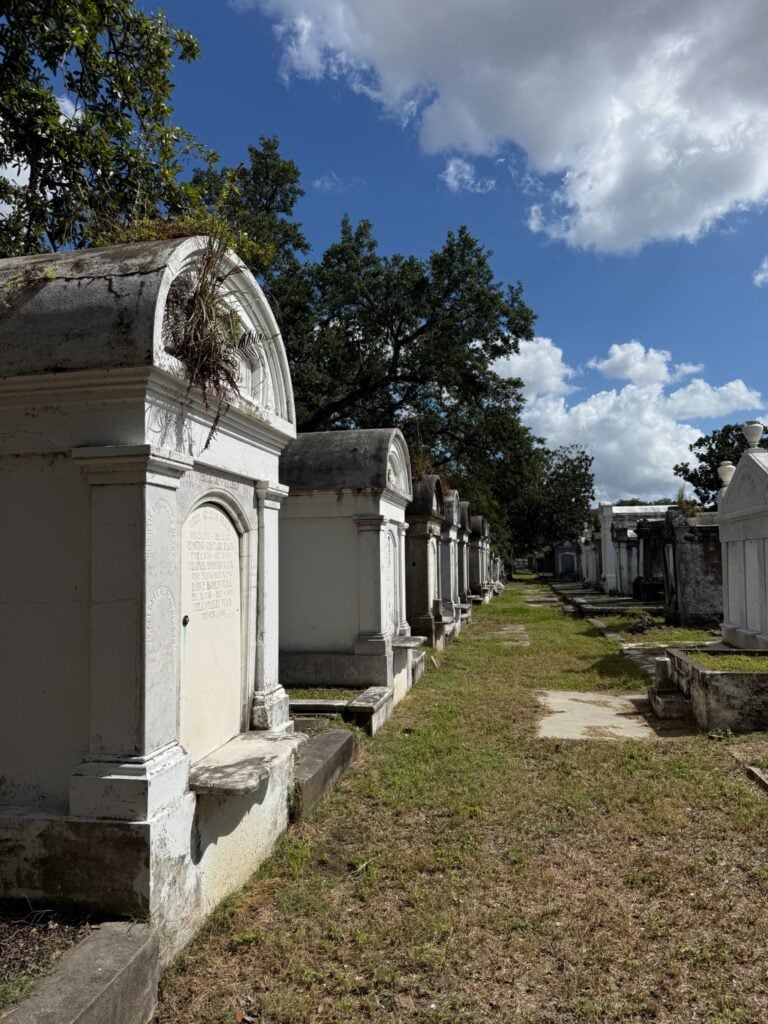 A row of weathered above-ground tombs in Lafayette Cemetery No. 1, with ornate arched fronts and carved memorial details. A grassy pathway runs between the tombs beneath mature trees and a bright blue sky with scattered clouds.