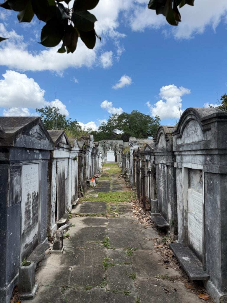 A narrow pathway through Lafayette Cemetery No. 1 lined with rows of weathered above-ground tombs and mausoleums. Cracked concrete walkways and patches of grass run between the tombs beneath mature trees and a bright blue sky with scattered clouds.