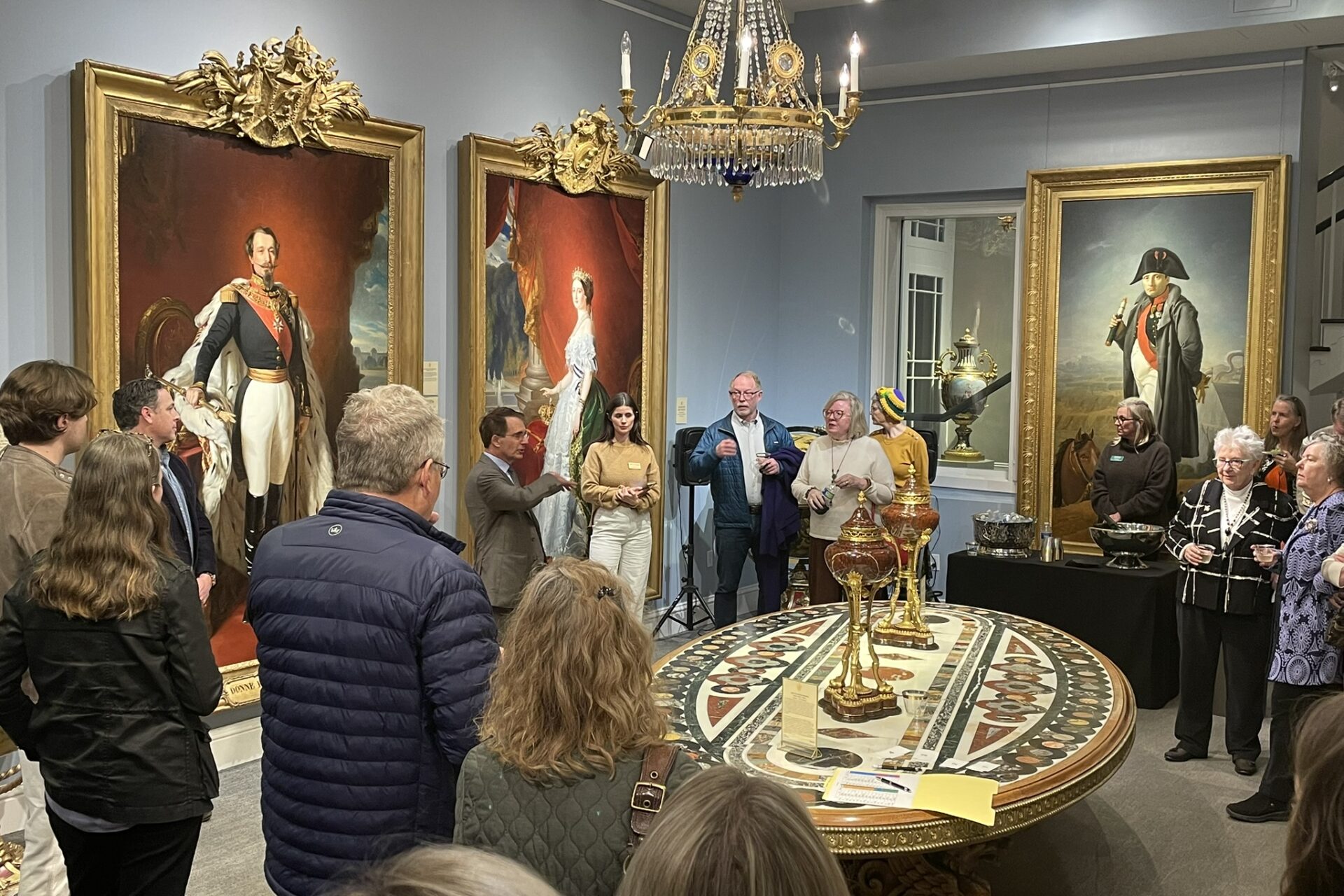 A group gathers for a guided talk inside an ornate museum gallery filled with large gilded portraits, decorative objects, an elaborate chandelier, and a richly patterned central table.