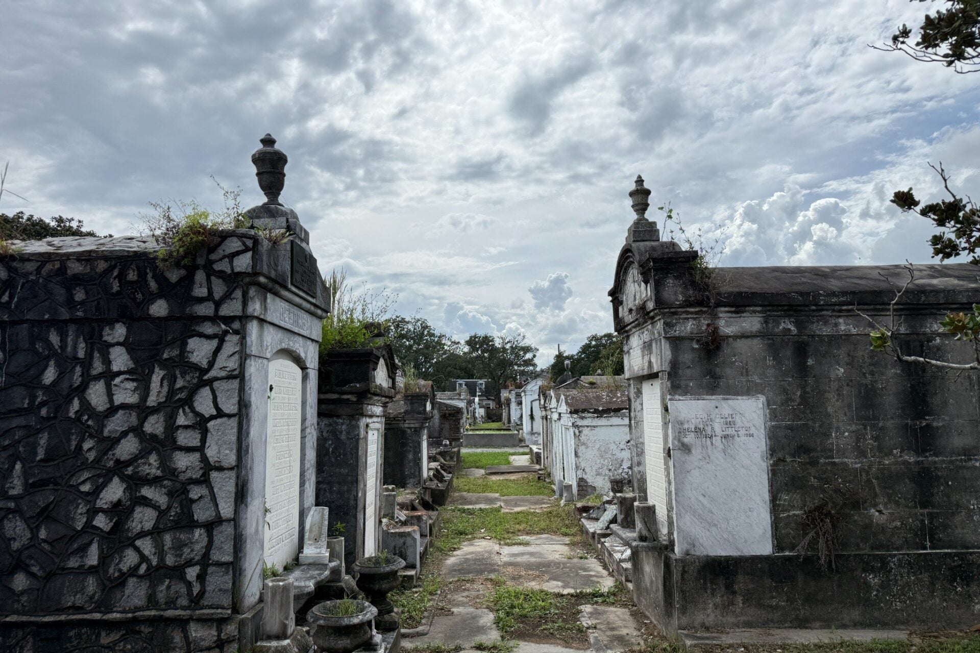 A narrow pathway lined with weathered above-ground tombs in Lafayette Cemetery No. 1, with ornate stone mausoleums, carved memorials, and urns surrounded by grass and vegetation beneath a cloudy sky.