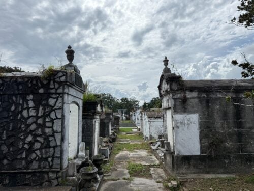 A narrow pathway lined with weathered above-ground tombs in Lafayette Cemetery No. 1, with ornate stone mausoleums, carved memorials, and urns surrounded by grass and vegetation beneath a cloudy sky.